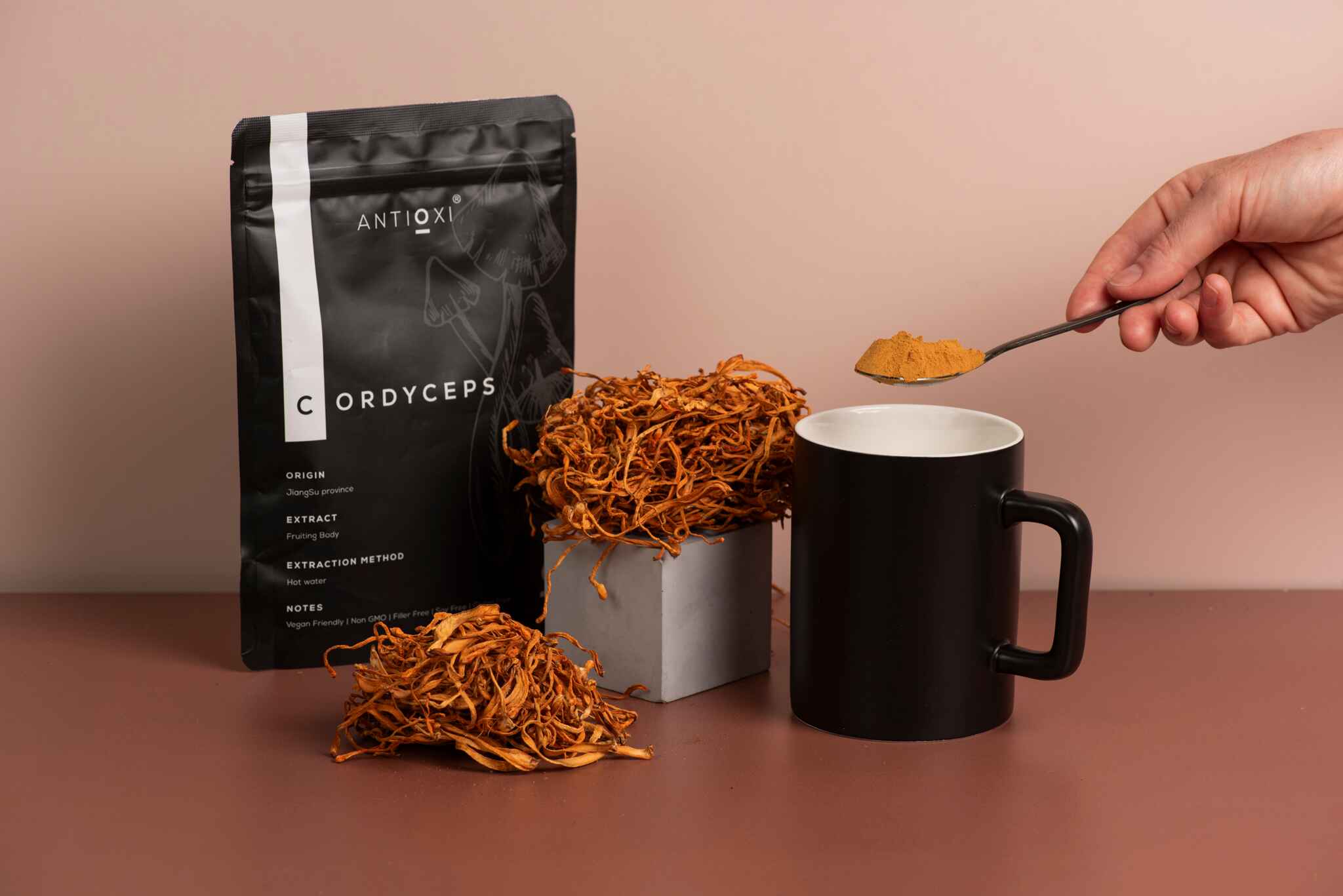 A black Antioxi Cordyceps pouch next to dried Cordyceps mushrooms and a black mug, with a hand holding a spoonful of powdered Cordyceps over the mug.