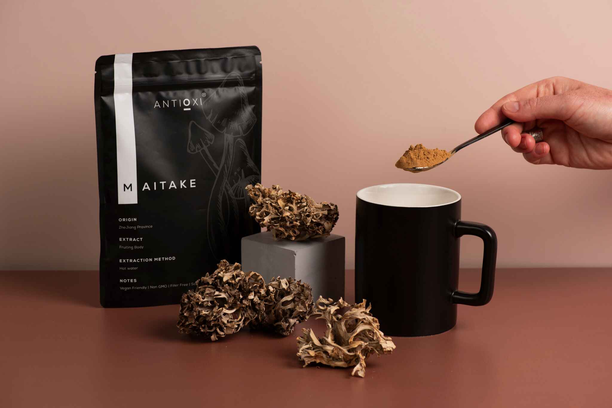 A black Antioxi Maitake mushroom supplements pouch next to dried Maitake mushrooms and a black mug, with a hand holding a spoonful of brown-orange, powdered Maitake over the mug.