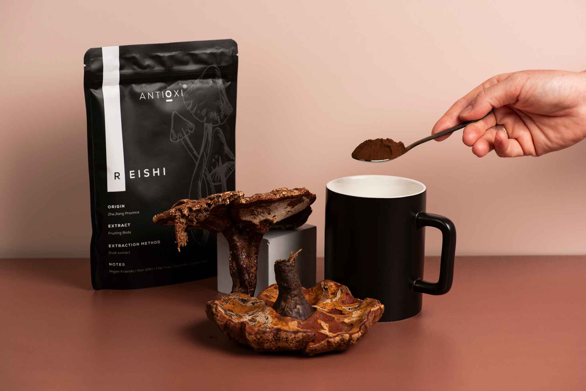A pouch of Antioxi Reishi mushroom supplements is displayed alongside whole Reishi mushrooms, with a hand holding a spoonful of Reishi powder over a black mug.