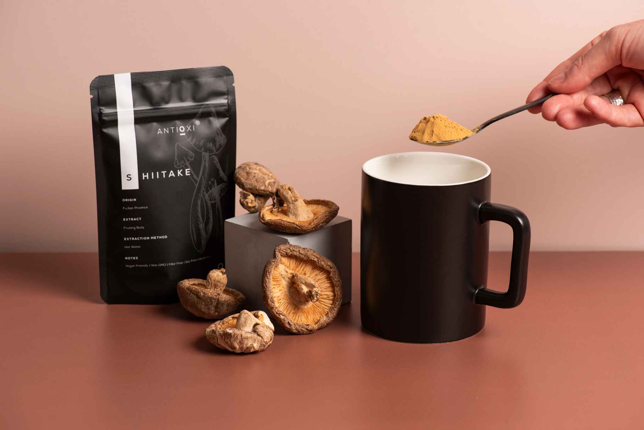 A black Antioxi Shiitake mushroom supplements pouch next to dried Shiitake mushrooms and a black mug, with a hand holding a spoonful of yellow, powdered Shittake over the mug.