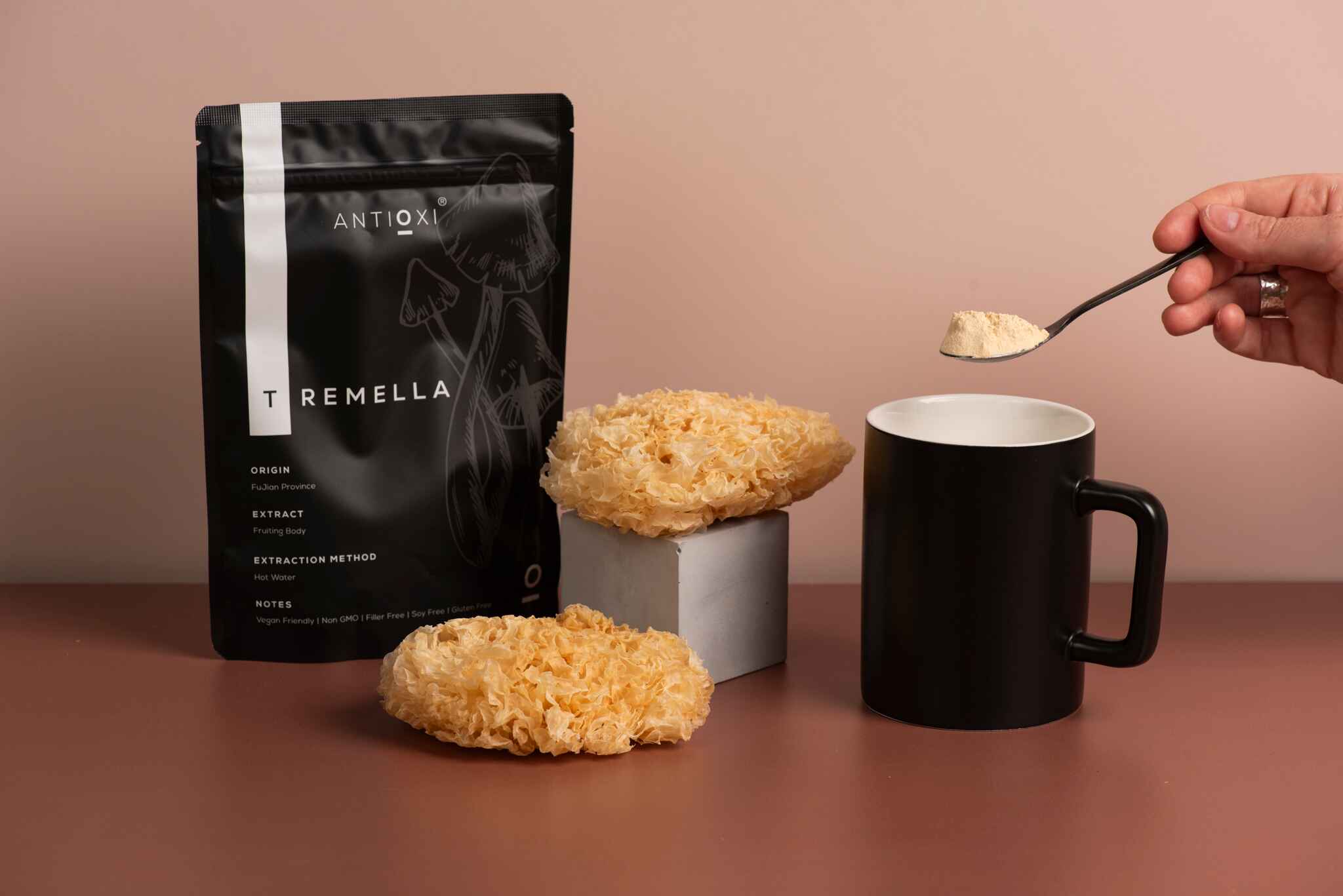 A black Antioxi Tremella mushroom supplements pouch next to dried Tremella mushrooms and a black mug, with a hand holding a spoonful of white, powdered Tremella over the mug.