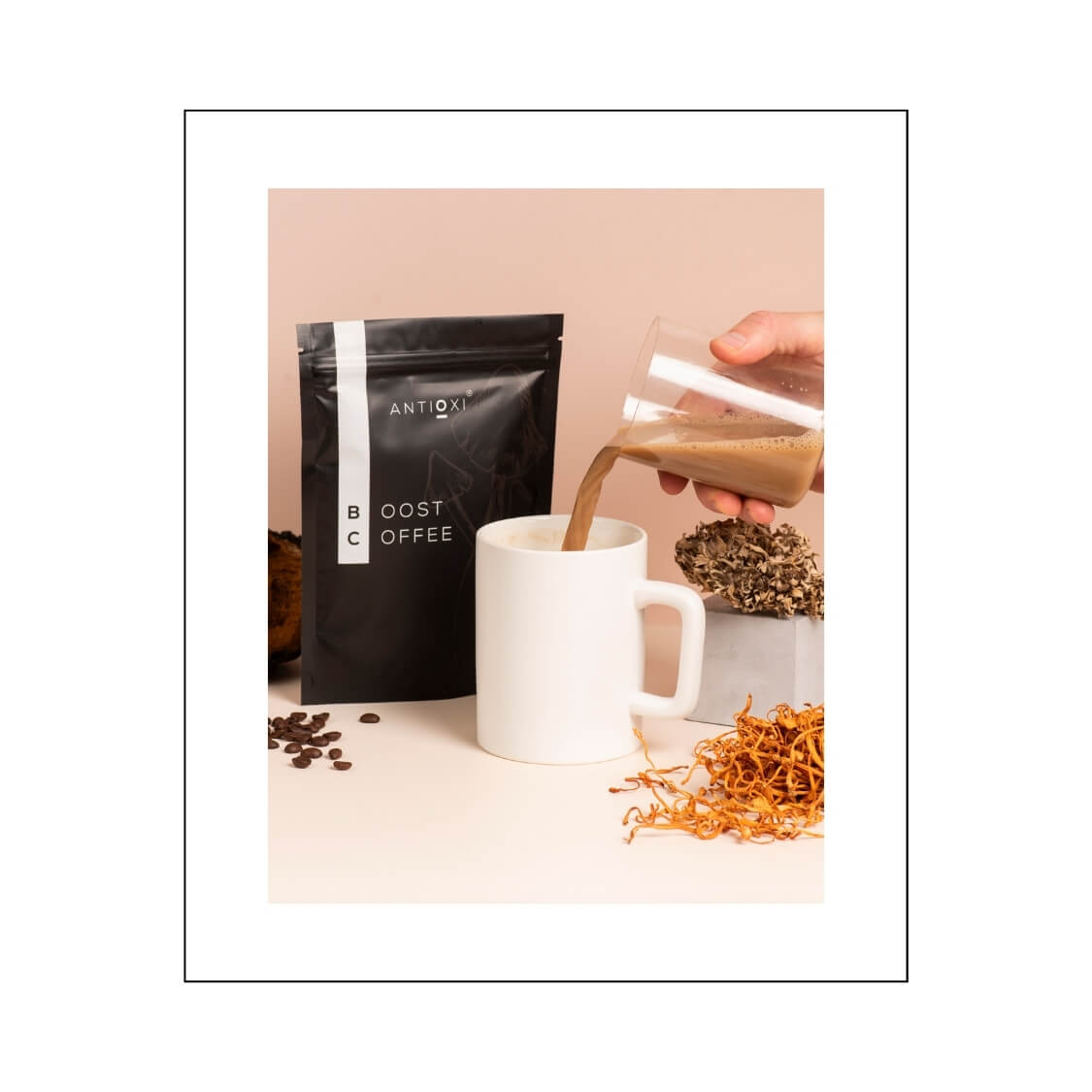 A pouch of Antioxi Boost Mushroom Coffee is displayed alongside a white mug, with coffee being poured into the mug. Surrounding the setup are scattered coffee beans and dried Cordyceps mushrooms.