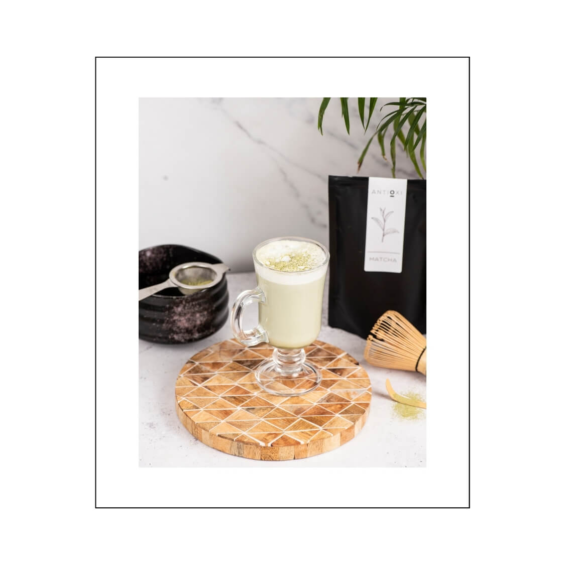 A cup of frothy Antioxi Matcha tea sits on a wooden board, with a whisk and a packet of Antioxi Matcha beside it.