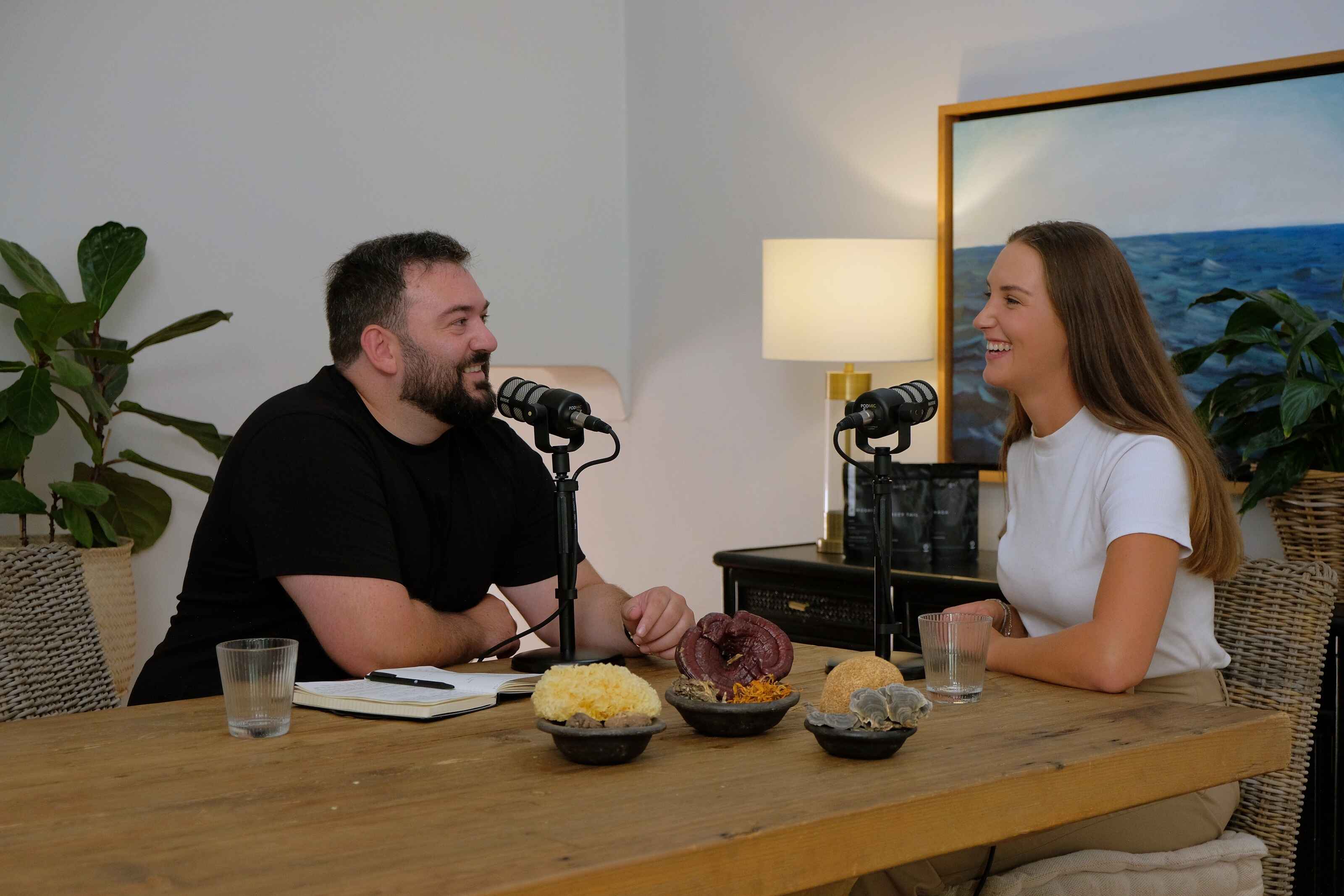 Carmen, one of the guests on the What's Wrong With You (WWWY) podcast, is seated at a wooden table, engaged in a lively conversation with Antioxi co-founder Marko Grensemann. The table is adorned with bowls containing various mushrooms, emphasizing the podcast's focus on health.
