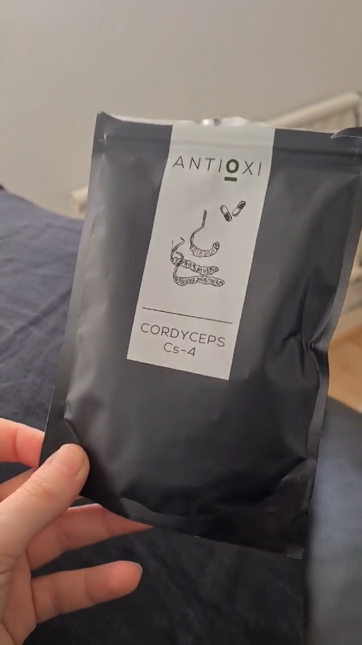 Close-up of a hand holding an Antioxi Cordyceps mushroom supplement pouch. The pouch is black with a white label that features an illustration of Cordyceps mushrooms and two capsules.