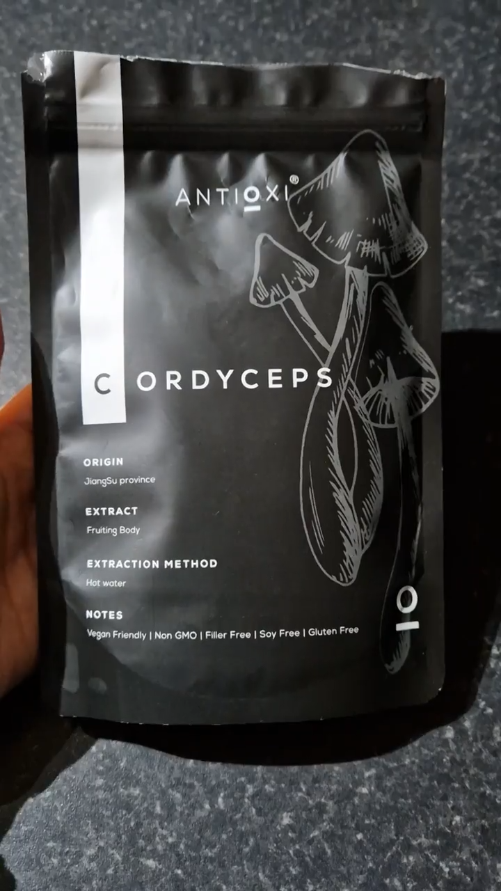 A close-up of a hand holding an Antioxi Cordyceps mushroom supplement pouch. The black pouch features the Antioxi logo, an illustration of Cordyceps mushrooms, and notes on its vegan-friendly, non-GMO, and filler-free attributes.